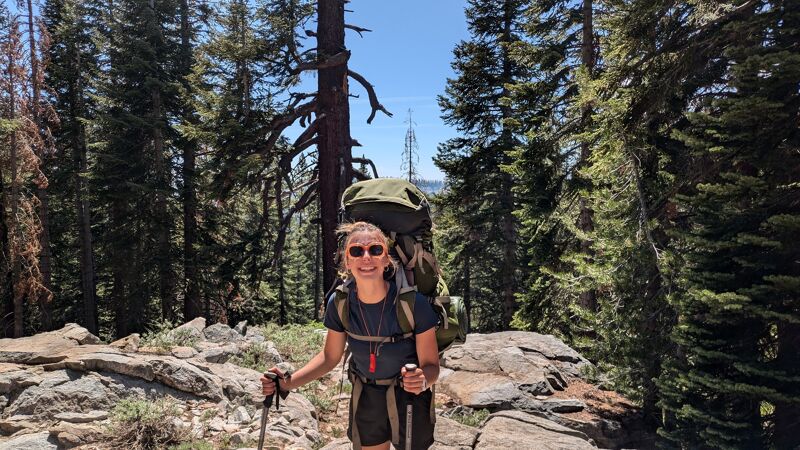 A woman with sunglasses and a large backpack is hiking in a forest. She is holding hiking poles and smiling at the camera. The forest is dense with tall trees and there are rocks and foliage on the ground. The sky is visible in the background.